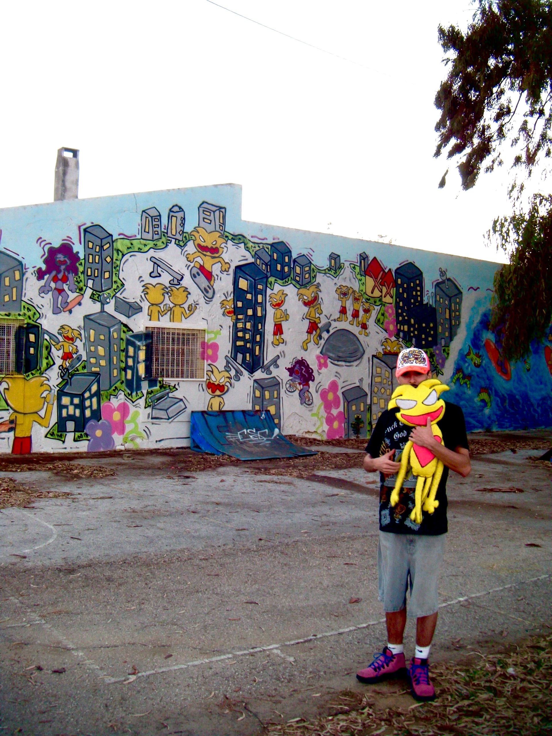 Mural Brincar p Sempre: prédios, flores e personagens amarelas numa parede, com o Tomás em frente a segurar um boneco